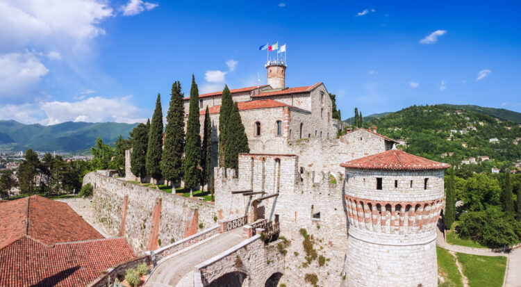 An aerial view of the historic Brescia castle, showing its turrets and drawbridge in the foreground and hills in the background. It is a sunny day and the flags are flying atop the highest turret.
