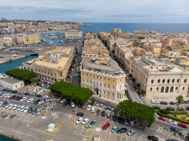 An aerial drone shot of the Grand Hotel in Ortigia, looking across to the sea and marina beyond, with cars parked along the edge.
