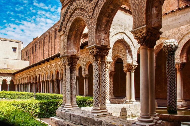 The intricately carved arches and columns of a cloister in Monreale Cathedral, Sicily. Sunlight illuminates the warm-toned stonework, creating patterns of light and shadow.