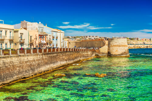 View of an embankment in Ortigia. Picturesque view of the historical part of Syracuse. Beautiful coastline with green-blue, transparent water of The Ionian Sea, Sicily, Italy