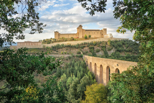 A fortress in Italy in Spoleto, Province of Perugia, stands on a hilltop, framed by lush green trees. A long, arched aqueduct stretches across the valley below, connecting the fortress to the surrounding landscape.