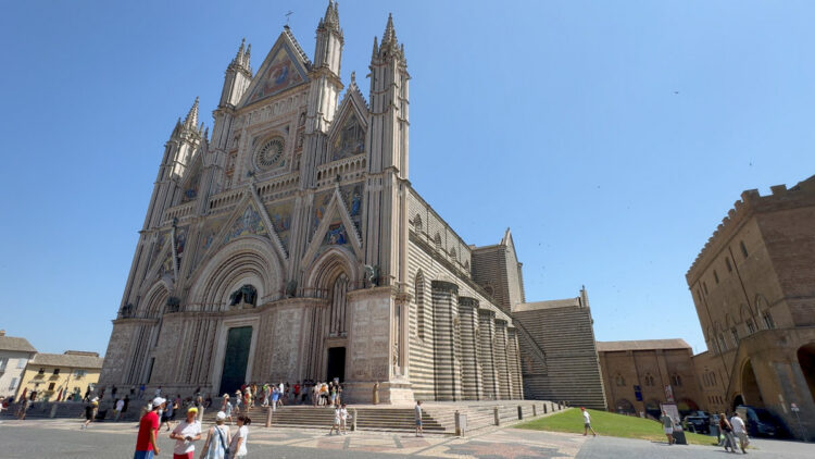 Orvieto Cathedral in central Italy, with an ornate striped stone façade, detailed mosaics, and visitors gathered in the square under a clear blue sky.