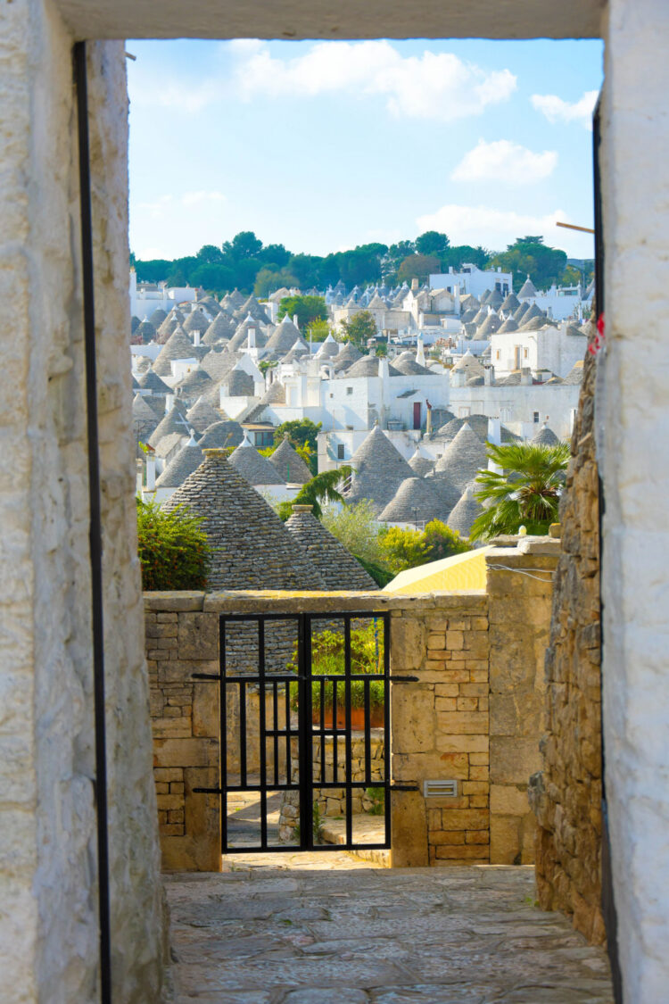 View of the trulli houses of Alberobello in southern Italy, with conical stone roofs seen through a stone doorway in bright daylight.